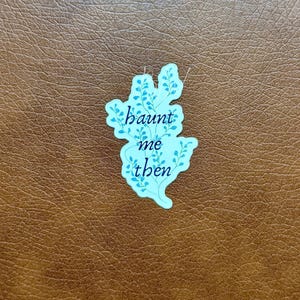 K&ouml;nnte beinhalten: Ein hellblauer Aufkleber mit den Worten "haunt me then" in einer Schreibschrift. Der Aufkleber hat die Form einer stilisierten Blume mit blauen Blattdetails. Der Aufkleber befindet sich auf einer braunen, lederartigen Oberfl&auml;che.
