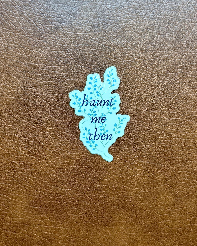 K&ouml;nnte beinhalten: Ein hellblauer Aufkleber mit den Worten "haunt me then" in einer Schreibschrift. Der Aufkleber hat die Form einer stilisierten Blume mit blauen Blattdetails. Der Aufkleber befindet sich auf einer braunen, lederartigen Oberfl&auml;che.