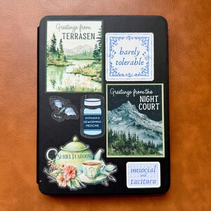 May include: A black e-reader with several colorful stickers on it. The stickers feature illustrations of a forest, a teacup, a bottle, a bat, and text that reads "Greetings from TERRASEN", "barely tolerable", "Greetings from the NIGHT COURT", "HYPAXIA'S DEWORMING MEDICINE", "SURIEL TEAROOM", and "unsocial and taciturn".