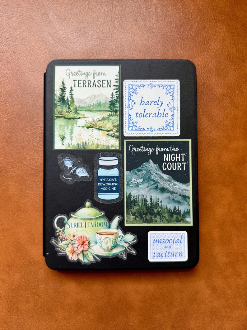 May include: A black e-reader with several colorful stickers on it. The stickers feature illustrations of a forest, a teacup, a bottle, a bat, and text that reads "Greetings from TERRASEN", "barely tolerable", "Greetings from the NIGHT COURT", "HYPAXIA'S DEWORMING MEDICINE", "SURIEL TEAROOM", and "unsocial and taciturn".