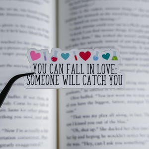 May include: A clear sticker with the text "YOU CAN FALL IN LOVE: SOMEONE WILL CATCH YOU" in black. The sticker has pink, red, and teal hearts, and colorful science beakers. The sticker is held by black tweezers, with a book in the background.