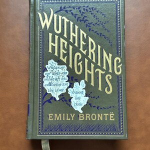 K&ouml;nnte beinhalten: Ein gebundenes Buch mit dem Titel "Wuthering Heights" von Emily Bront&euml;. Das Cover ist olivgr&uuml;n mit goldfarbenen Buchstaben und dunkelblauen dekorativen Elementen. Das Buch hat ein Leseband und Zitat-Aufkleber.