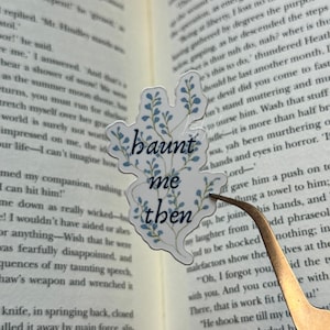 K&ouml;nnte beinhalten: Ein transparenter Aufkleber mit dem Text "haunt me then" in einer stilisierten Schriftart, verziert mit blauen floralen Elementen. Der Aufkleber befindet sich auf einem offenen Buch, das von einer Pinzette gehalten wird, was auf ein Lesezeichen hindeutet.