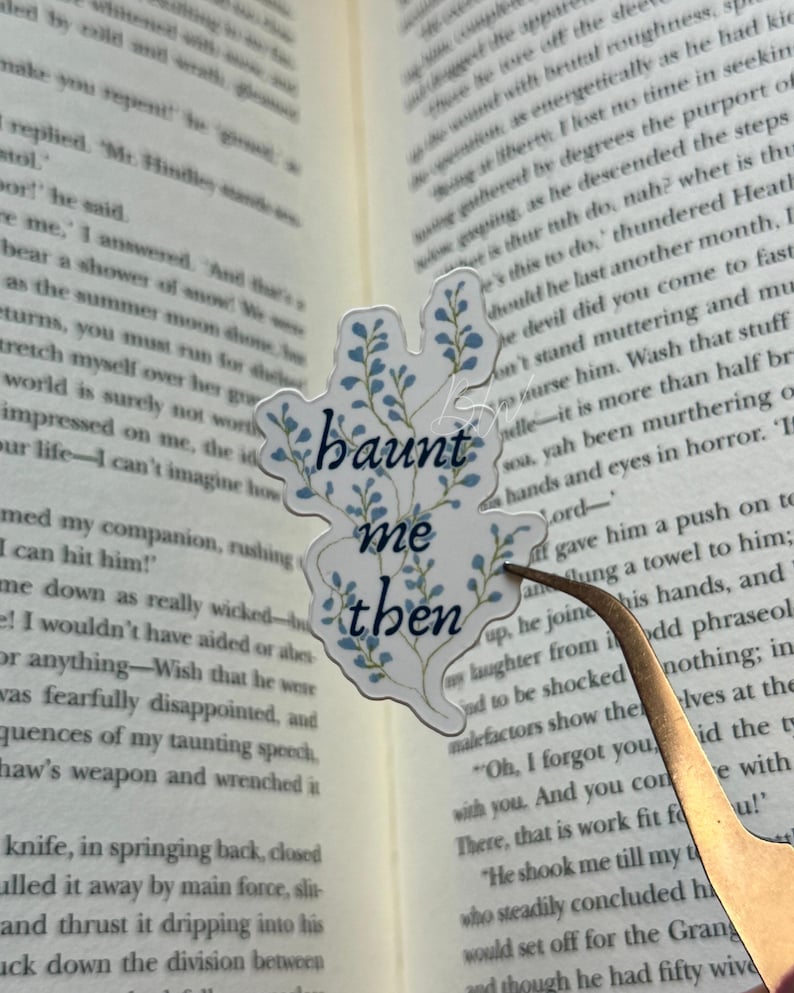 K&ouml;nnte beinhalten: Ein transparenter Aufkleber mit dem Text "haunt me then" in einer stilisierten Schriftart, verziert mit blauen floralen Elementen. Der Aufkleber befindet sich auf einem offenen Buch, das von einer Pinzette gehalten wird, was auf ein Lesezeichen hindeutet.