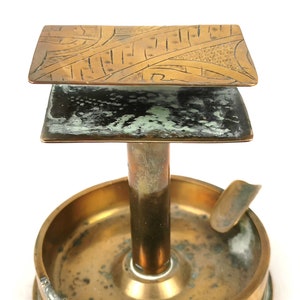 May include: A brass ashtray with a decorative top and a built-in cigarette rest. The top features an intricate geometric design.