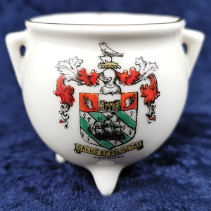 May include: White ceramic pot with three legs, featuring a coat of arms with a ship, a castle, and a bird. The text "Salus et Felicitas Torquay" is printed below the coat of arms.