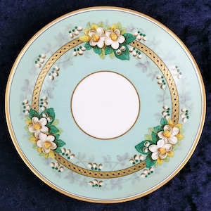 May include: A light blue saucer with a gold rim and a white center. The saucer is decorated with a floral pattern of white flowers and green leaves.