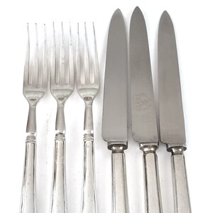 May include: A set of six silver-colored flatware pieces, including three forks and three knives. The forks have four tines, and the knives have a rounded tip. Each piece has a decorative handle with an emblem.