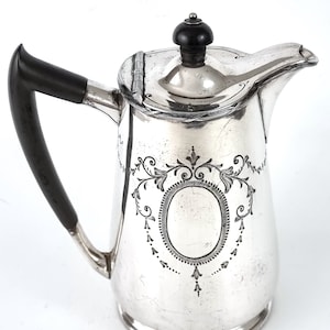 Puede incluir: Una cafetera de plata con asa negra y un diseño grabado decorativo. La cafetera tiene una tapa con un pomo negro.
