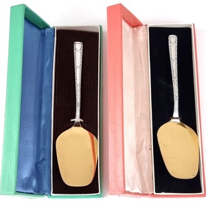 Op de afbeelding: Twee zilveren serveerscheppen met sierlijke handvatten worden getoond in hun originele dozen. De dozen zijn gevoerd met zwart fluweel en hebben een groene en roze buitenkant.