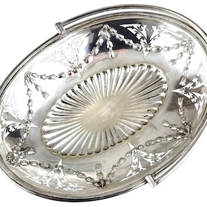 Puede incluir: Un plato de servir ovalado de plata con un diseño floral decorativo y un asa. El plato tiene un centro elevado y un acabado liso y pulido.