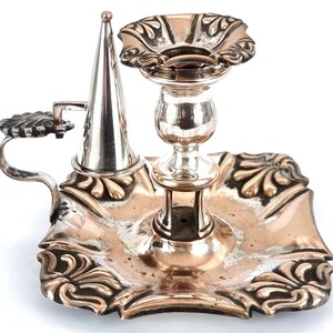 Puede incluir: Un candelero de plata y cobre con una base decorativa y un apagador. El candelero tiene una base redonda con un borde elevado y un tallo central que sostiene un portavelas en forma de copa. El apagador es un pequeño cono puntiagudo que se encuentra en un brazo articulado.