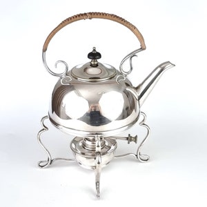 May include: Silver teapot with a wicker handle and a burner stand. The teapot has a polished finish and a round body.