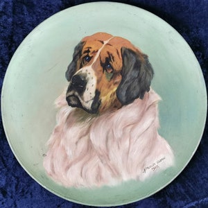 May include: A round, light green plate featuring a painted portrait of a Saint Bernard dog. The dog has brown and white fur with black accents and a pinkish-white fluffy neck. Signed "H. Morley George 1925."