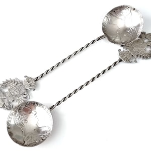 May include: A pair of silver spoons with twisted handles. Each spoon features a round bowl with a raised design of a woman's profile and a coat of arms. The spoons are connected by a twisted handle.