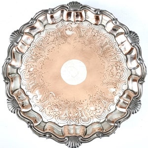 Può includere: Un vassoio da portata in argento con un bordo a festoni e intricati motivi floreali incisi. Il vassoio ha un centro rialzato e una finitura lucida.