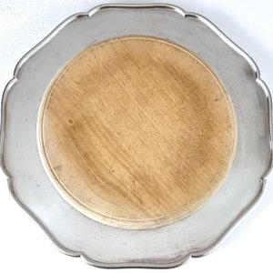 Puede incluir: Una bandeja de servir plateada con un inserto de madera para tabla de cortar. La bandeja tiene un borde festoneado y un acabado liso y pulido.