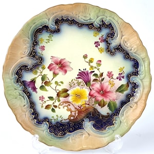 Puede incluir: Un plato de porcelana vintage con un diseño floral decorativo. El plato presenta un borde azul y dorado con una imagen central de flores rosas, amarillas y blancas.