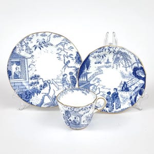 Könnte beinhalten: Ein blau-weißes Porzellan-Teeservice mit einer Tasse und zwei Tellern. Das Set hat einen goldenen Rand und ein detailliertes asiatisch inspiriertes Landschaftsdesign. Die Teller sind rund mit gewellten Rändern.