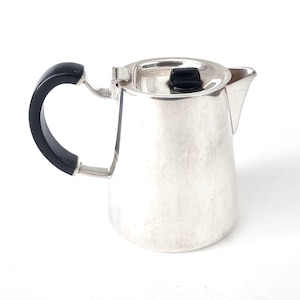 Puede incluir: Una cafetera plateada con asa y pomo de tapa negros. La cafetera tiene un asa curvada y un pico vertedor. La tapa con bisagras tiene un pequeño pomo negro. Probablemente hecha de acero inoxidable.