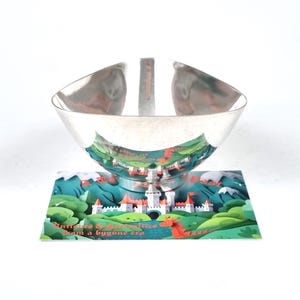 May include: A polished silver-toned bowl with a curved, four-sided rim, resting on a rectangular card featuring a colorful illustration of a castle, green hills, and a red dragon with the text Antiques & Curiosities from a bygone era.