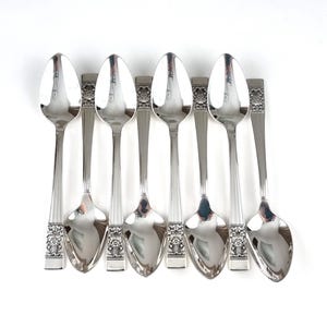 Könnte beinhalten: Sechs silberne Löffel, vertikal vor weißem Hintergrund angeordnet. Die Löffel haben ein klassisches Design mit einem dekorativen Blumenmuster am Griff und einer reflektierenden, ovalen Schale. Sie bestehen aus glänzendem, poliertem Metall.