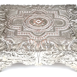 May include: A silver square cake stand with an ornate design. The stand has four legs and a raised center section with a floral pattern.