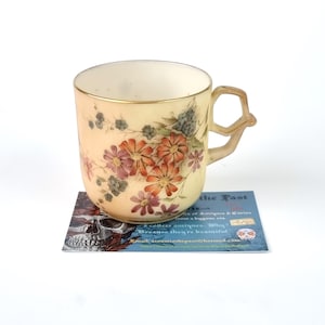 Puede incluir: Una taza de té vintage con un diseño floral en tonos naranja, rosa y morado. La taza tiene un asa única y un borde dorado. La taza está sobre una tarjeta con texto y una imagen de calavera.