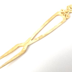 May include: A white bone hair fork with an ornate design at the top. The design features a crown and other intricate details.