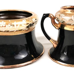 May include: A black and gold ceramic sugar bowl and creamer set with a floral design. The set has a gold rim and a gold band around the middle.