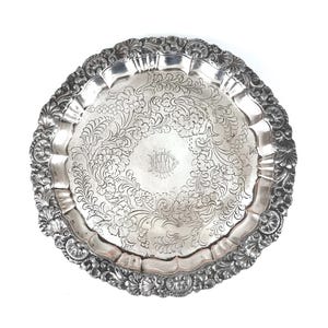 May include: A silver serving tray with an ornate floral design. The tray has a scalloped edge and a detailed border. The center of the tray features an engraved monogram. The tray is round and appears to be made of metal.