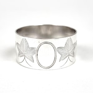 May include: A silver napkin ring with engraved maple leaf designs and an oval cartouche. The ring is cylindrical and made of a shiny, reflective metal. The leaves are detailed with etched lines.