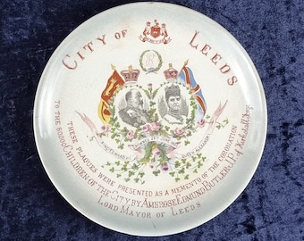 Puede incluir: Un plato de cerámica blanco con un diseño rojo y dorado que conmemora la coronación del rey Eduardo VII y la reina Alejandra en 1902. El plato tiene el texto "City of Leeds" y "Estas placas fueron presentadas como recuerdo de la coronación de los 80.000 niños de la ciudad por Ambrose Edmund Butler, Lord Mayor de Leeds."