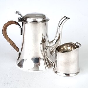 May include: A silver teapot with a brown wicker handle and a matching silver creamer. The teapot has a lid and a spout. The creamer has a handle and a spout.