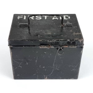 Puede incluir: Una caja de primeros auxilios de metal negro con tapa con bisagras y asa de transporte. La caja tiene una inscripción blanca que dice "FIRST AID".