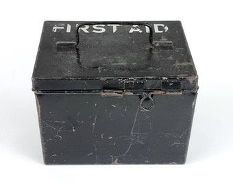 Op de afbeelding: Een zwarte metalen EHBO-doos met een scharnierend deksel en een draaggreep. De doos heeft een witte inscriptie met de tekst "FIRST AID".