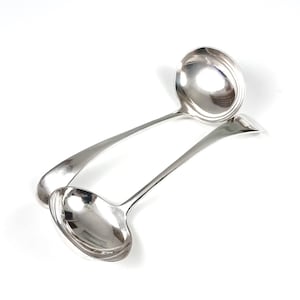 May include: Two silver-colored ladles with long, curved handles and rounded bowls are displayed against a white background. The ladles are positioned at a slight angle, with one overlapping the other. The reflective surfaces of the ladles create highlights.