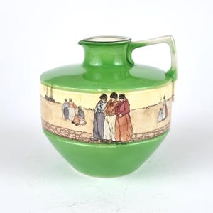 May include: A green ceramic jug with a handle, featuring a painted scene of people walking on a path. The jug has a cream-coloured band with the scene painted on it.