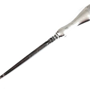 May include: A silver shoe horn with a hook on one end and a rounded handle with an engraved inscription. The handle is polished and the hook is tarnished.