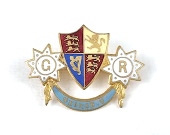 Puede incluir: Un broche de esmalte vintage con un escudo de armas, estrellas con las letras "G" y "R", y una pancarta azul claro con la inscripción "GEORGE V". El broche presenta detalles dorados y un escudo de armas.