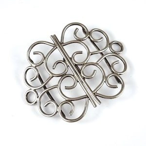 May include: Silver filigree clasp featuring a decorative design of interlocking swirls and loops. The clasp is open and ready to be attached to a garment or accessory.