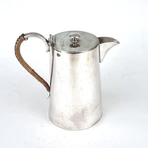 May include: A silver pitcher with a brown leather handle and a lid. The pitcher has a rounded body and a spout.