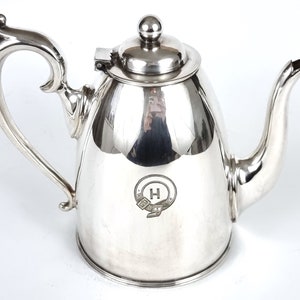 Peut inclure: Une théière en argent avec un corps rond et une poignée incurvée. La théière a un couvercle avec un bouton sur le dessus et un bec verseur sur le côté. La théière a une lettre "H" gravée à l'intérieur d'un cercle sur le devant.