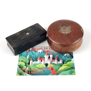 May include: Two antique wooden boxes. One is rectangular and black with a star design. The other is round and brown with a metal emblem. A colorful card with a castle illustration is also present.