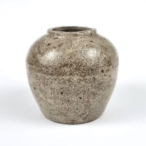 May include: A small, round ceramic vase with a textured, speckled surface. The vase is a muted grey colour with darker brown spots. The opening is small, and the overall shape is bulbous. This decorative vase is suitable for home decor.