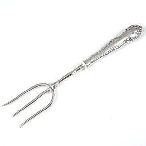 May include: A silver-toned metal serving fork featuring three long, slender tines and a decorative handle with intricate embossed scrollwork and rope-like detailing along the edges, terminating in a rounded, ornate finial at the top end.