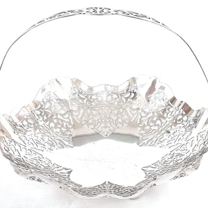 Könnte beinhalten: Ein silberfarbener, verzierter Korb mit Henkel. Der Korb hat einen gewellten Rand und aufwendige durchbrochene Muster im gesamten Korb und Henkel. Der Henkel ist gewölbt und an den Seiten des Korbes befestigt.