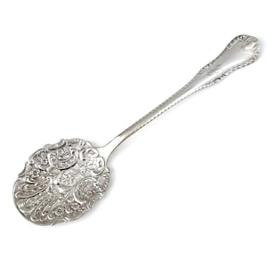 May include: A silver serving spoon with ornate floral and scrollwork detailing on the bowl. The handle has a delicate, engraved design. The spoon is likely vintage or antique, and is set against a white background.