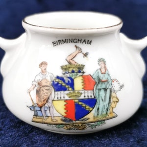 Peut inclure: Une tasse en céramique blanche à deux anses, ornée d'un blason coloré avec le texte "BIRMINGHAM" et "FORWARD" en bas.