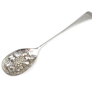 Puede incluir: Una cuchara de servir de plata con un diseño adornado de uvas y hojas. La cuchara tiene un mango largo y delgado y un cuenco redondeado.